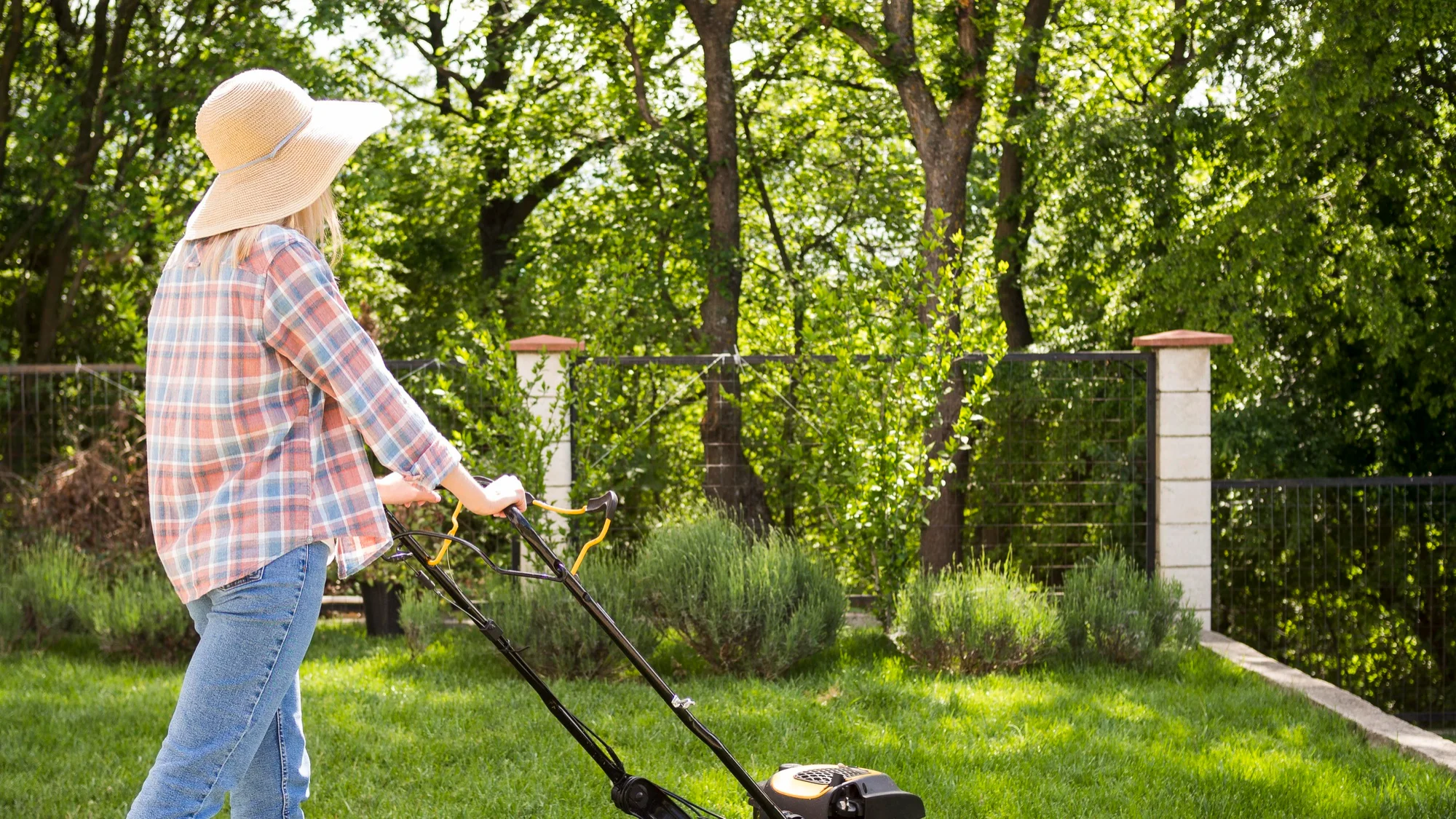 A woman in a sunhat mows the lawn in a lush backyard garden.