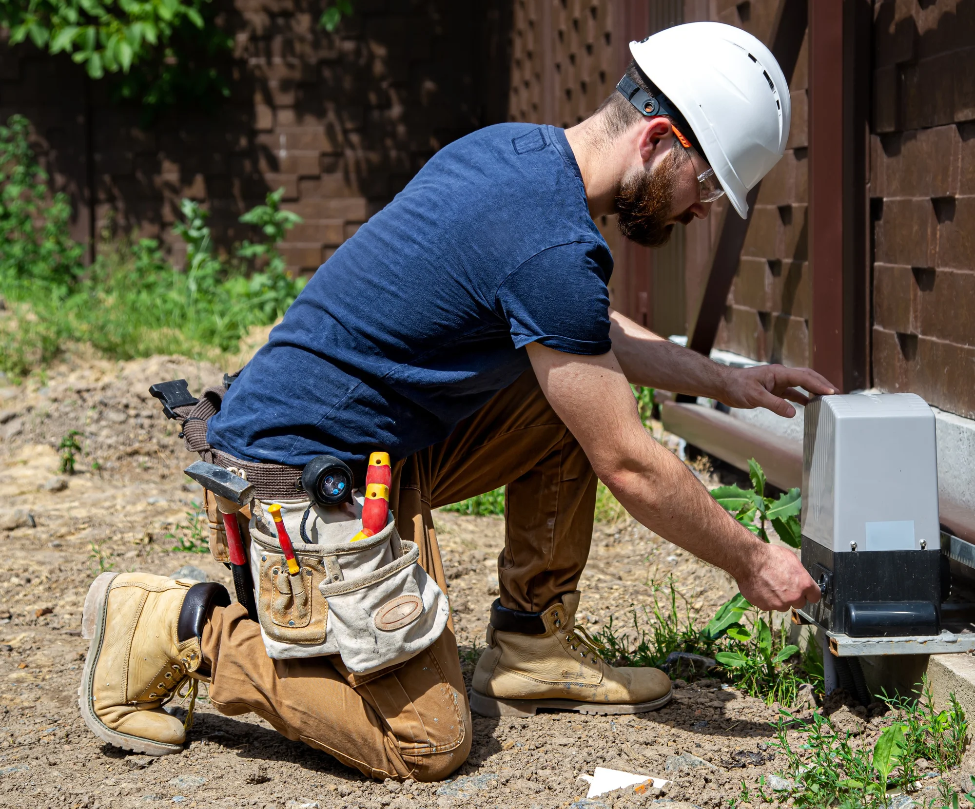 A construction worker kneels while inspecting outdoor equipment.